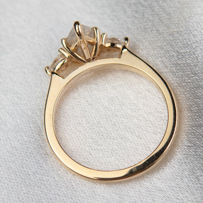 A three stone ring with a central pear-shaped diamond set in gold , flanked by two smaller pear diamonds, reflecting light on a soft, light, blurred background. 