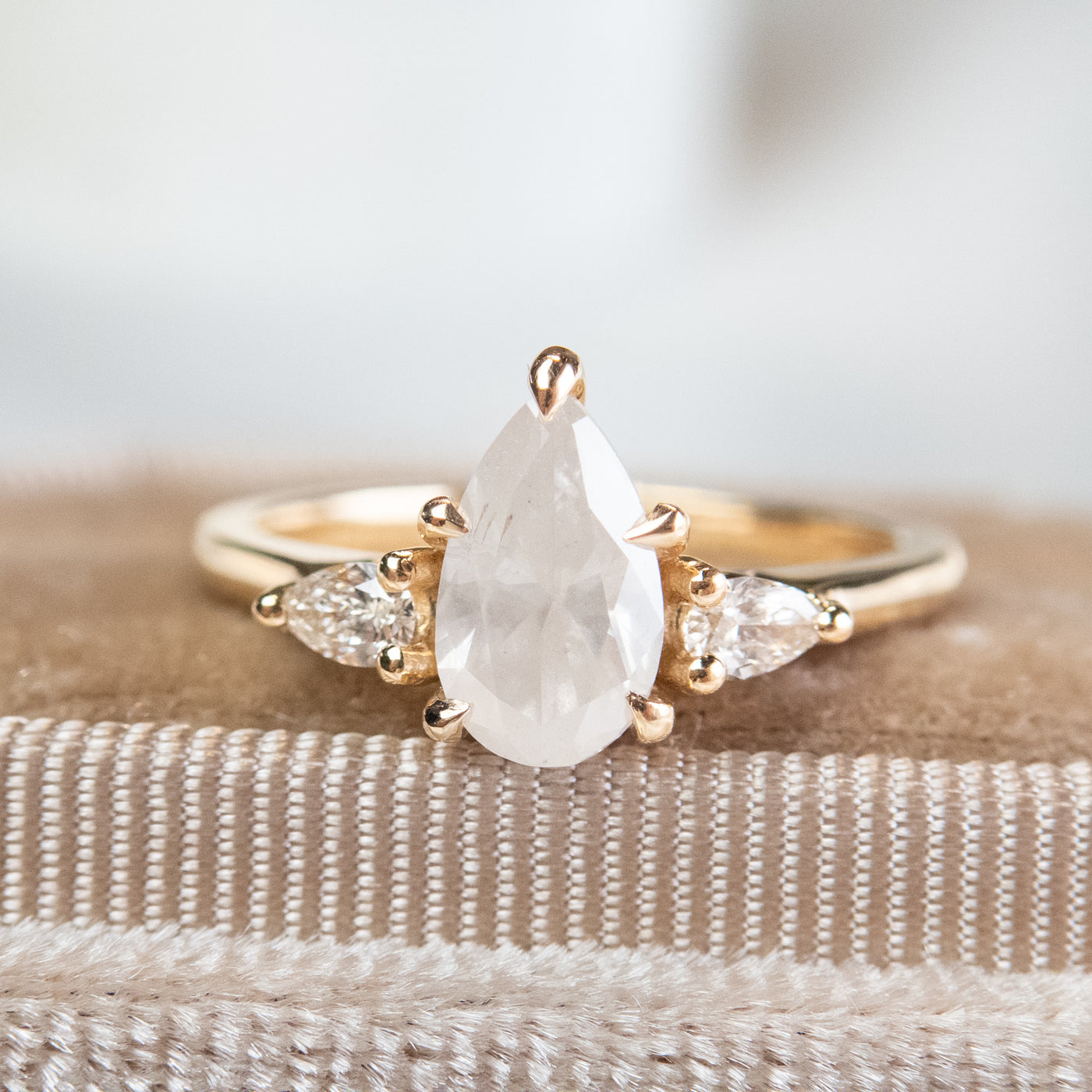 A three stone ring with a central pear-shaped fancy white diamond set in gold , flanked by two smaller pear diamonds, reflecting light on a soft, velvet ring box. 