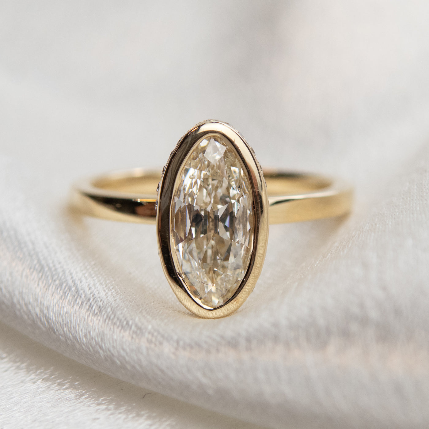 An elongated antique marquise-cut diamond ring bezel set with a polished gold band sits elegantly on a shiny white fabric. 