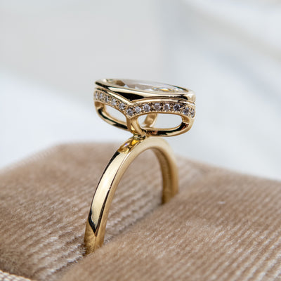 A side view of an elongated marquise-cut diamond ring bezel set with a polished gold band sits elegantly on a soft, velvet ring box. 