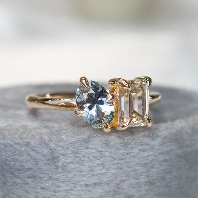 A stunning gold ring featuring an oval blue sapphire and a rectangular champange diamond, set elegantly against a soft grey fabric background.