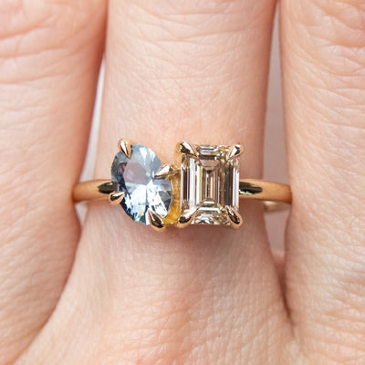 A stunning gold ring featuring an oval blue sapphire and a rectangular Champagne diamond worn on a finger. 