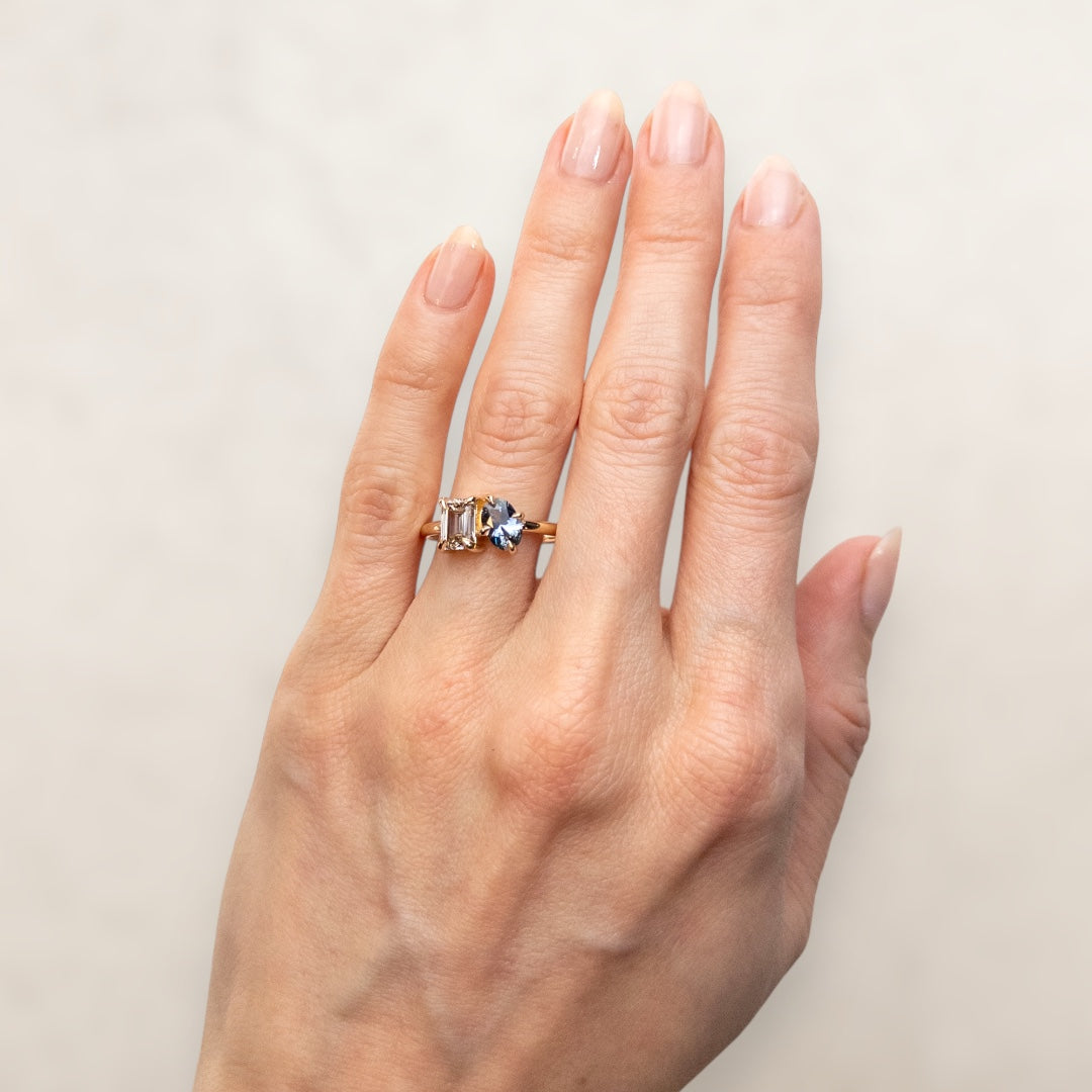 A stunning gold ring featuring an oval blue sapphire and a rectangular Champagne diamond worn on a hand.