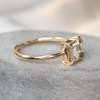 A stunning gold ring featuring an oval blue sapphire and a rectangular champange diamond, set elegantly against a soft grey fabric background.