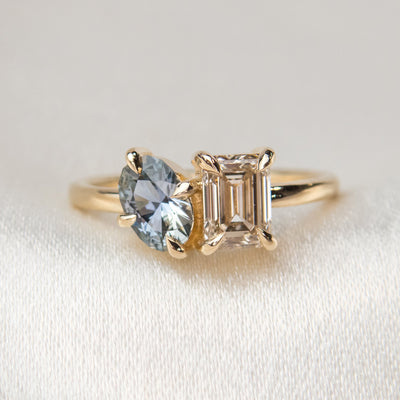 A stunning gold ring featuring an oval blue sapphire and a rectangular champange diamond, set elegantly against a soft fabric background.