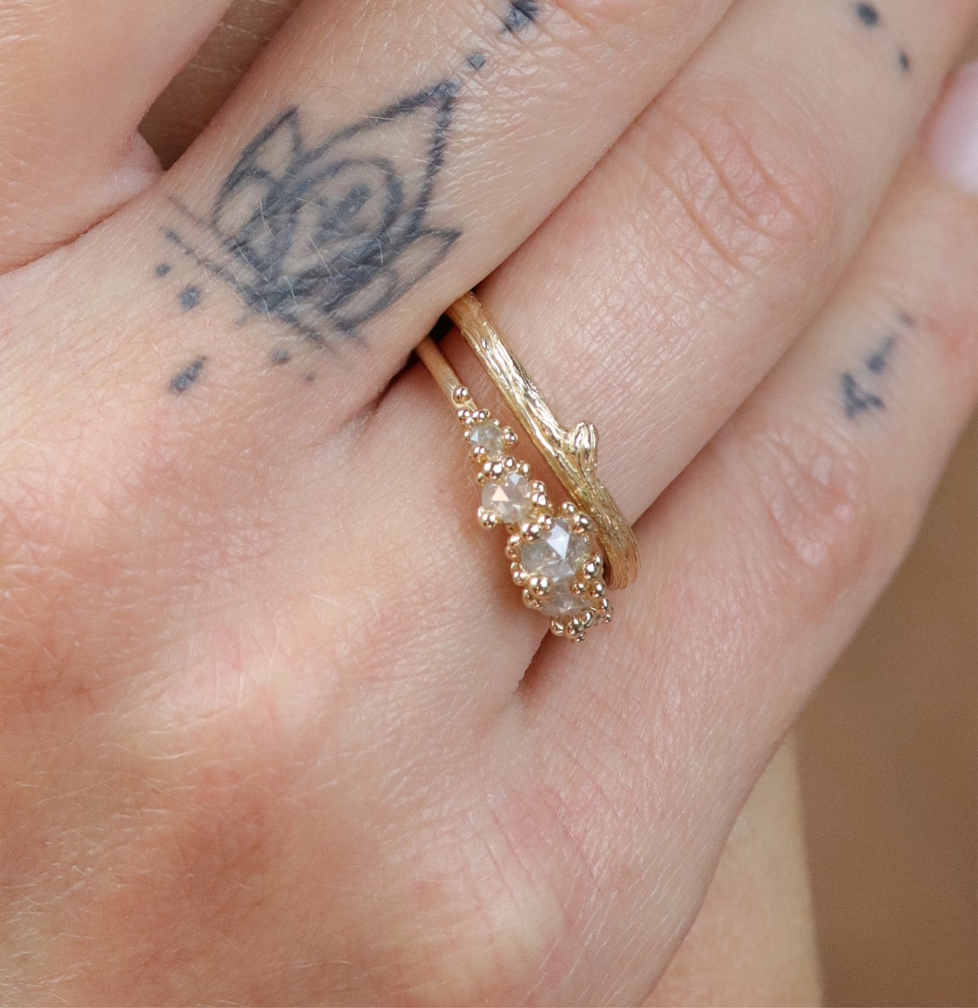 A delicate gold ring with a textured, nature-inspired design resembling a twisted twig stacked on a tattooed hand with a diamond rosecut ring.