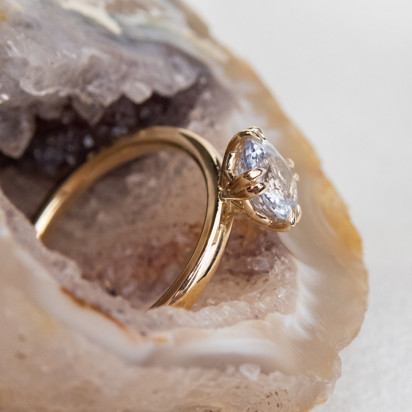 A side profile view of a gold solitaire ring with a colourless oval brilliant sapphire set in double claw prongs, set against natural crystal. 