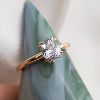 A gold solitaire ring with a colourless oval brilliant sapphire set in double claw prongs, set atop a blue-green ring holder. 