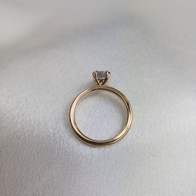 A side profile view of a gold solitaire ring with a colourless oval brilliant sapphire set in double claw prongs, set against a light fabric backdrop. 