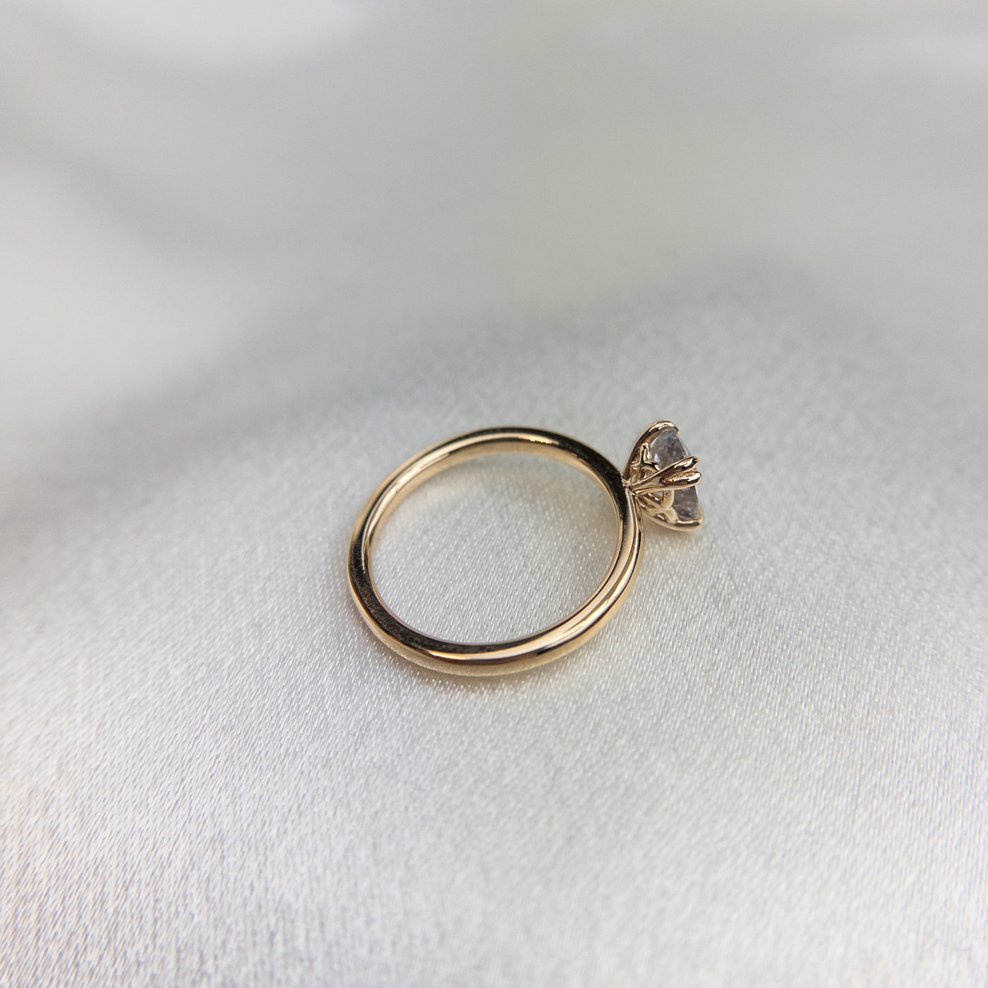 A side profile view of a gold solitaire ring with a colourless oval brilliant sapphire set in double claw prongs, set against a light fabric backdrop. 