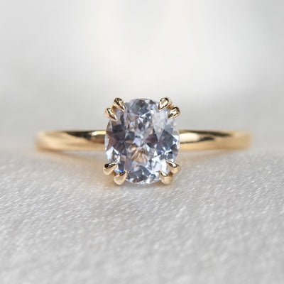 A gold solitaire ring with a colourless oval brilliant sapphire set in double claw prongs, set against a light fabric backdrop. 