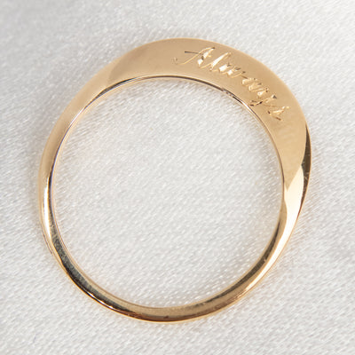 A stunning gold ring with a meaningful quote saying always, shown laying flat on white fabric from above.