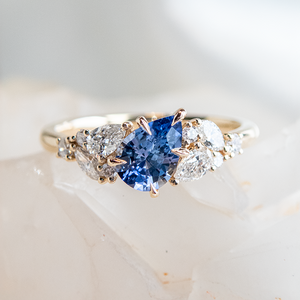 A close up photo of a gold engagement ring with a blue pear shaped gemstone surrounded by clusters of diamonds in different shapes on each side, resting on a white crystal.