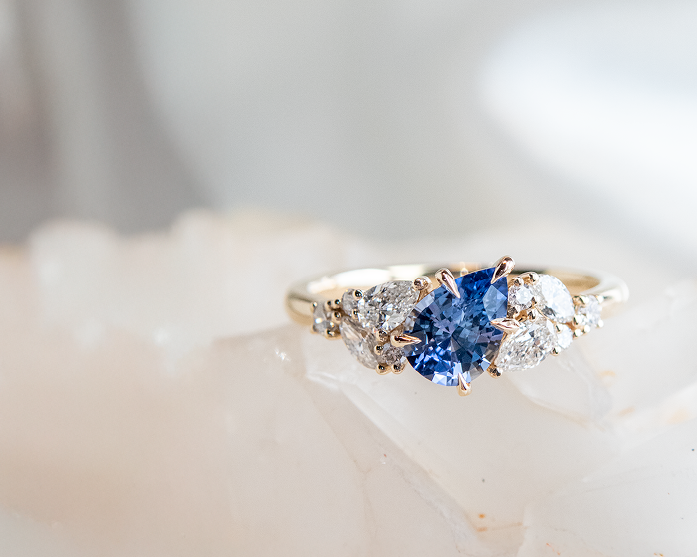 A gold engagement ring with a blue pear shaped gemstone surrounded by clusters of diamonds in different shapes on each side, resting on a white crystal.