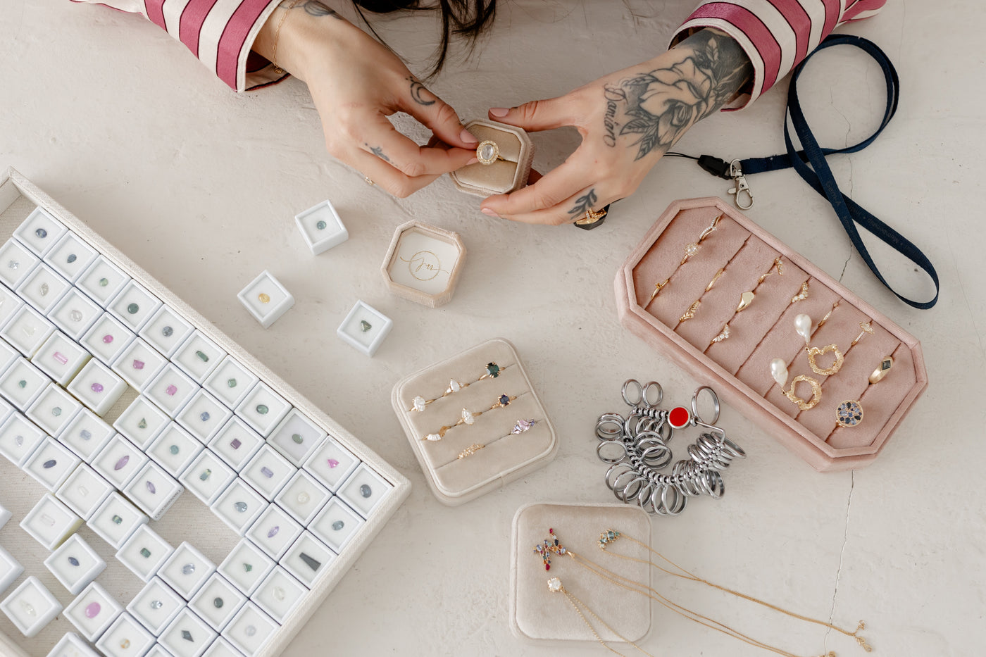 shot taken from above showing a woman holding a diamond ring in a jewelry box, surrounding her is a tray of fine gold jewelry like rings, earrings and pendants and a tray full of pastel colored gemstones in small white individual boxes.