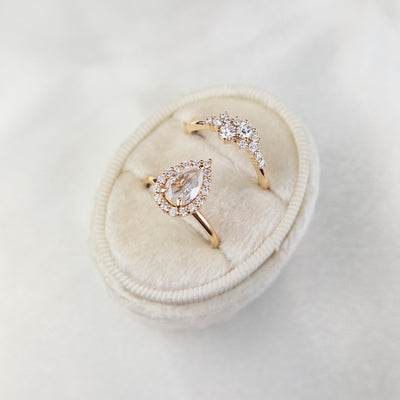 A stunning gold ring featuring a cluster of sparkling diamonds, set in a graduated design. Shown here in a cream ring box with a pear shaped milky diamond halo engagement ring. 
