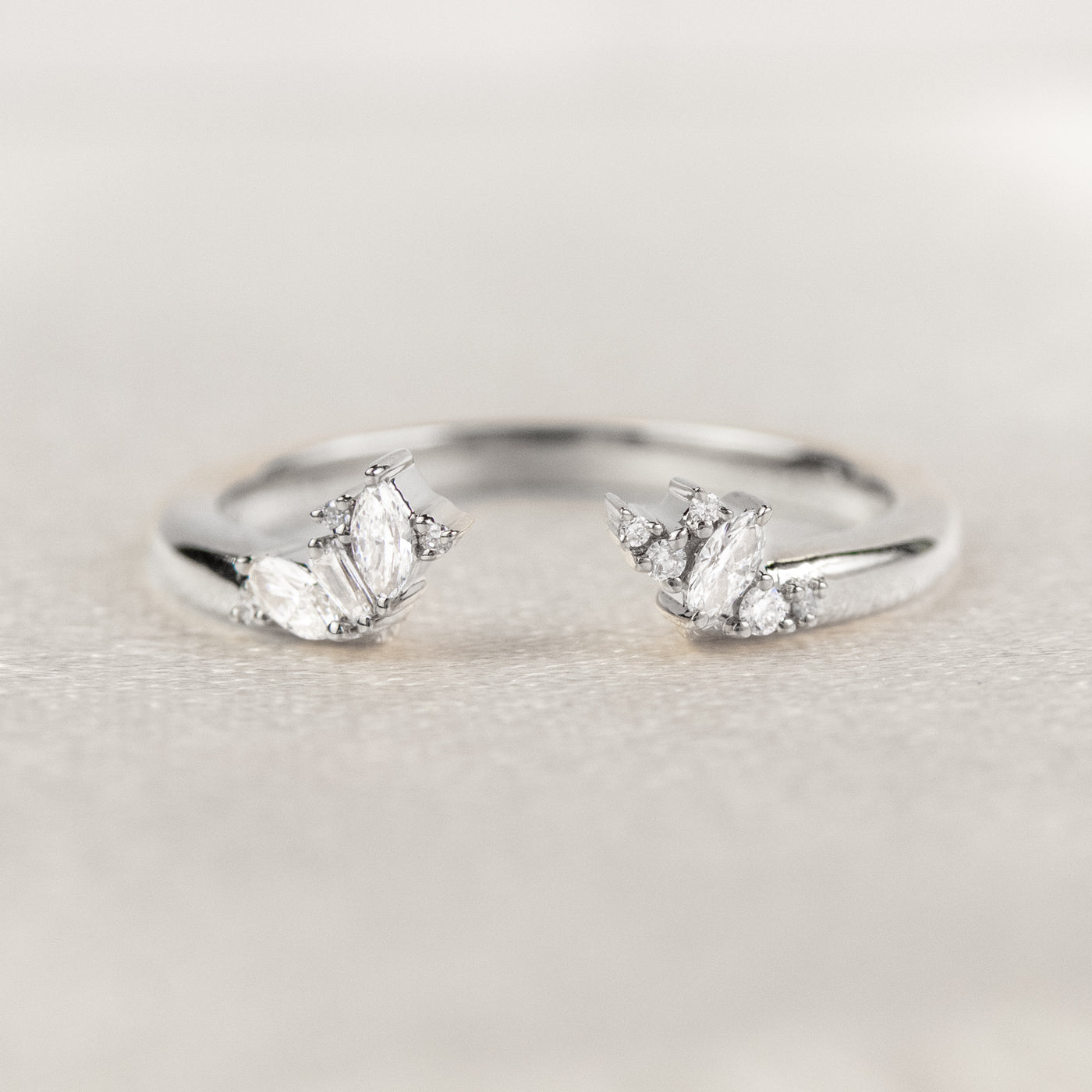 White gold open-ended ring with marquise and round cut diamonds on each end, stacked with an engagement ring on a soft-focus light background.
