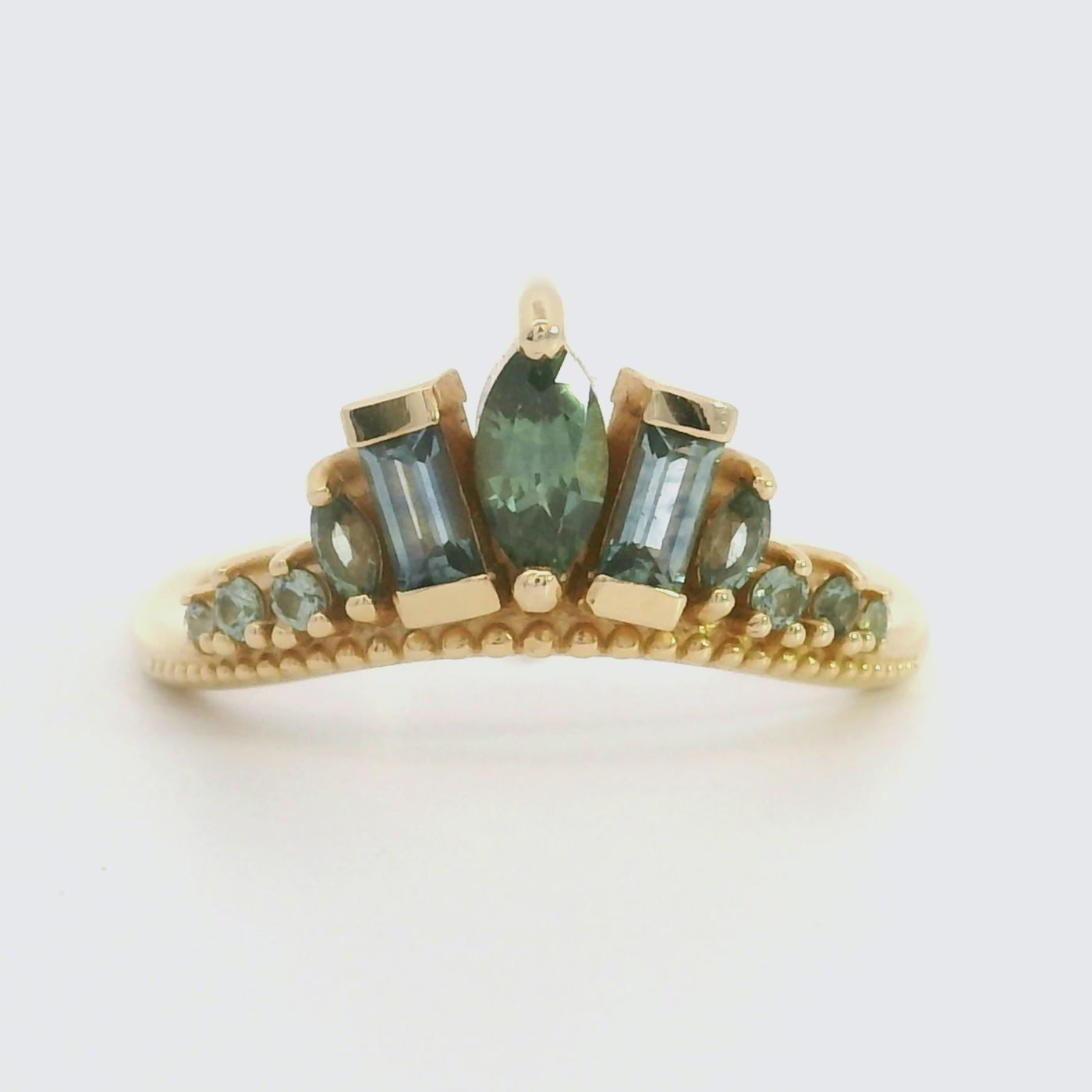 A video of a delicate gold ring adorned with blue and teal sapphires, featuring a central marquise stone and two rectangular stones in a crown design, reflecting light beautifully.