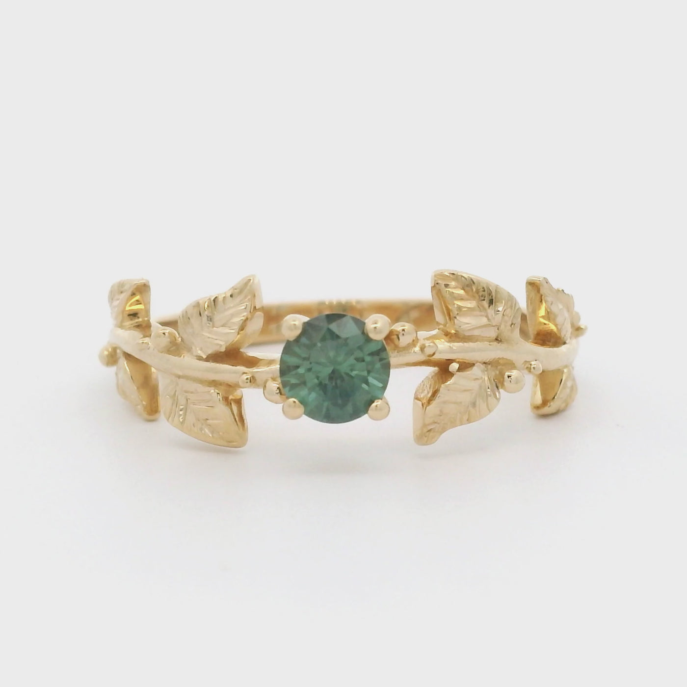 180 degree video of a nature-inspired gold ring with a round brilliant teal sapphire, featuring intricate leaf design band. 