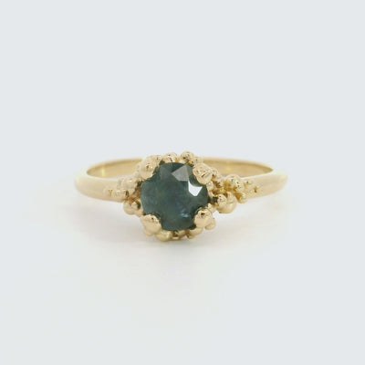 A gold ring with a textured band features a round brilliant cut blue sapphire, set in ornate gold prongs, on a white background. 