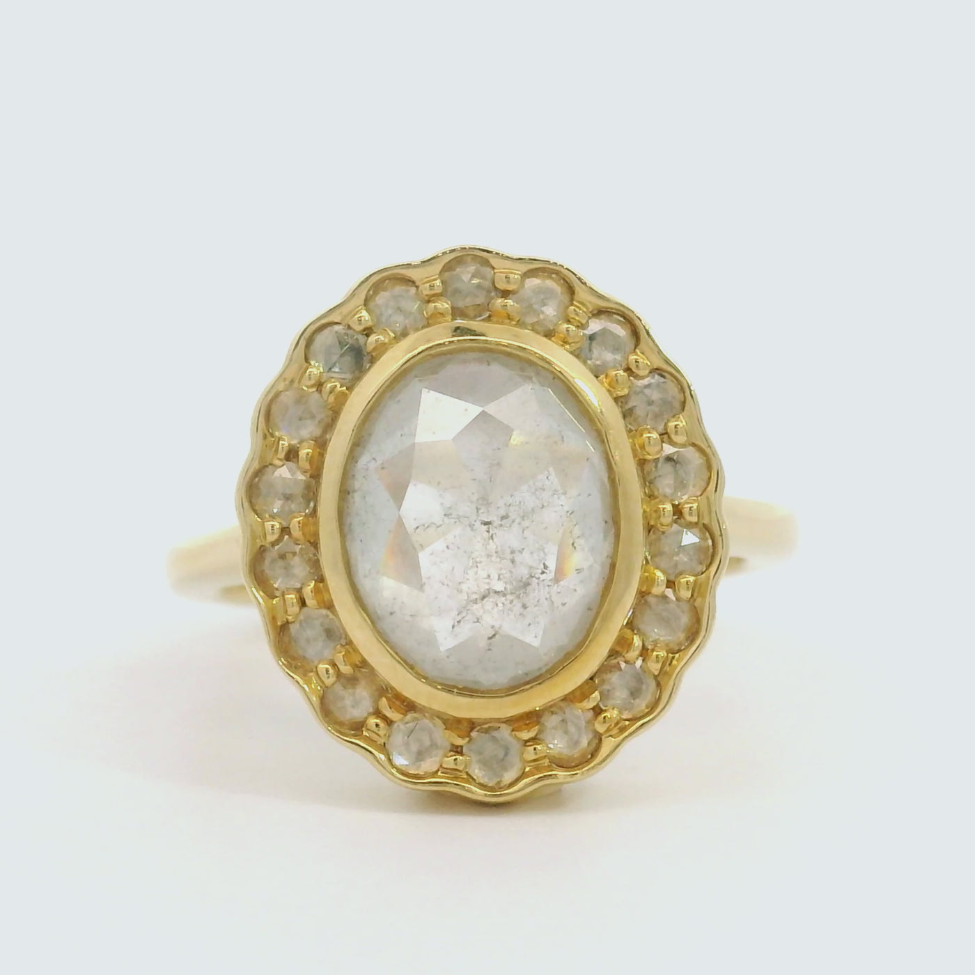 A gold ring with an oval, translucent grey salt and pepper diamond at its center is surrounded by a halo of small round rose cut diamonds, on a white background. 