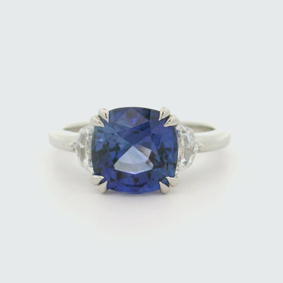 A 180 degree video of a large cushion shaped blue sapphire ring flanked by white cadillac cut diamonds set in a classic platinum setting, shown on a reflective white background. 