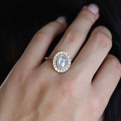 A gold ring with an oval, translucent grey salt and pepper diamond at its center is surrounded by a halo of small round rose cut diamonds, worn on a hand.