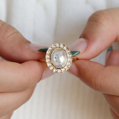 A gold ring with an oval, translucent grey salt and pepper diamond at its center is surrounded by a halo of small round rose cut diamonds, held by two hands against a soft, neutral background.