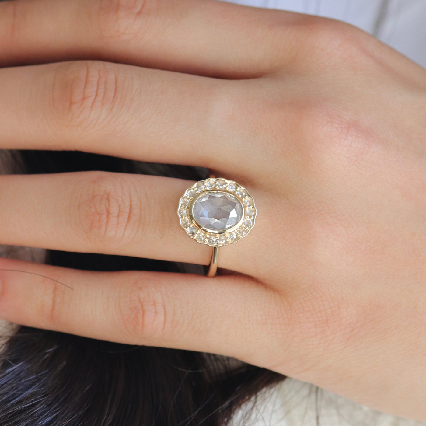 A gold ring with an oval, translucent grey salt and pepper diamond at its center is surrounded by a halo of small round rose cut diamonds, worn on a hand.