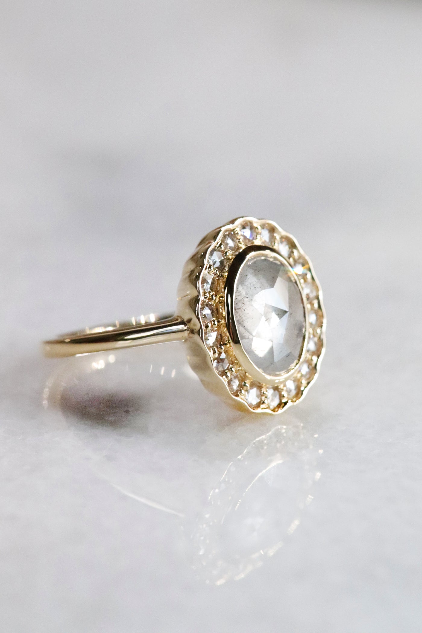 A gold ring with an oval, translucent grey salt and pepper diamond at its center is surrounded by a halo of small round rose cut diamonds, set against a soft, neutral background.