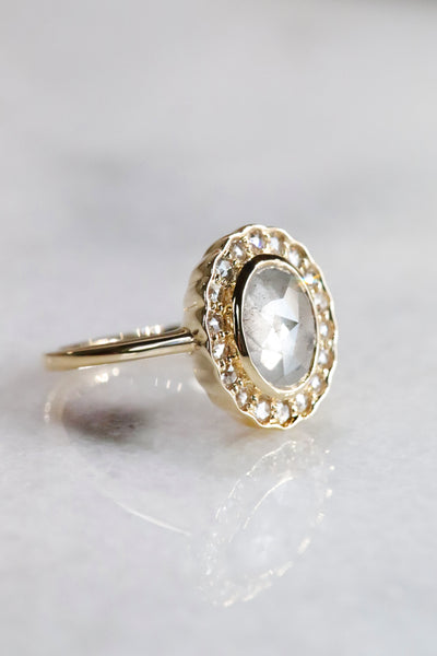 A gold ring with an oval, translucent grey salt and pepper diamond at its center is surrounded by a halo of small round rose cut diamonds, set against a soft, neutral background.