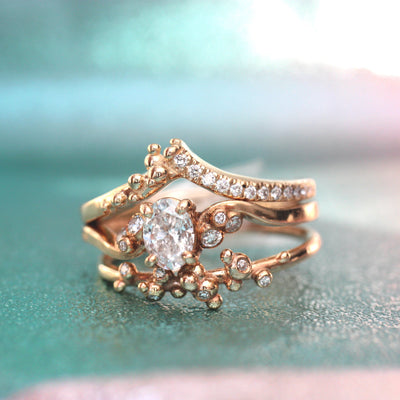A delicate gold wedding set featuring an oval center diamond and sparkling accents, artistically entwined against a soft teal background.