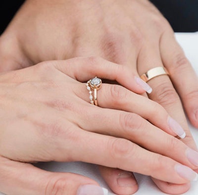 A woman rests her hand over a mans hand, each wearing their wedding and engagement rings. His is a flat gold simple band and hers is a diamond solitaire with small diamond details.