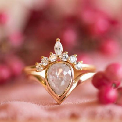 A delicate gold ring featuring a pear-shaped diamond centerpiece, crowned with smaller sparkling stones, set against a soft pink background.