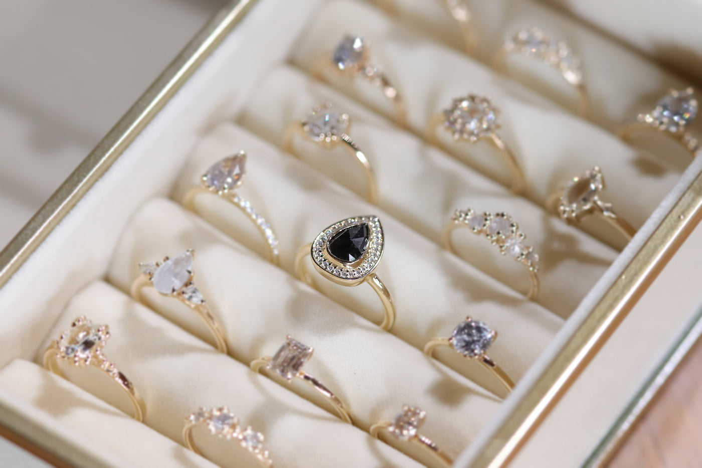 A jewelry box displays elegant gold rings with various gemstone shapes, including a distinct black pear shaped design, arranged neatly. 