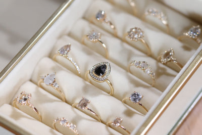 A jewelry box displays elegant gold rings with various gemstone shapes, including a distinct black pear shaped design, arranged neatly. 