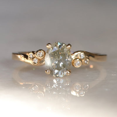 A gold ring with a fancy grey oval diamond at center, flanked by three small round diamonds on each side, lies on a soft white surface.