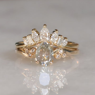 A gold ring with a fancy grey oval diamond at center, flanked by three small round diamonds on each side, stacked with an intricate crown-like diamond band lies on a soft white surface.