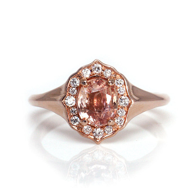 Elegant ring with an oval brilliant cut pink sapphire surrounded by a halo of small diamonds set in rose gold, against a white background. 