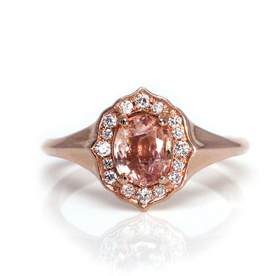 Elegant ring with an oval brilliant cut pink sapphire surrounded by a halo of small diamonds set in rose gold, against a white background. 