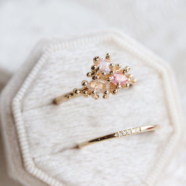 Simple and elegant gold stacking ring with rounded band and 5 pave diamonds, in a white velvet ring box with a engagement ring featuring a cluster of 3 unique pink toned sapphires.