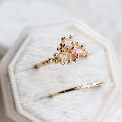 Simple and elegant gold stacking ring with rounded band and 5 pave diamonds, in a white velvet ring box with a engagement ring featuring a cluster of 3 unique pink toned sapphires.