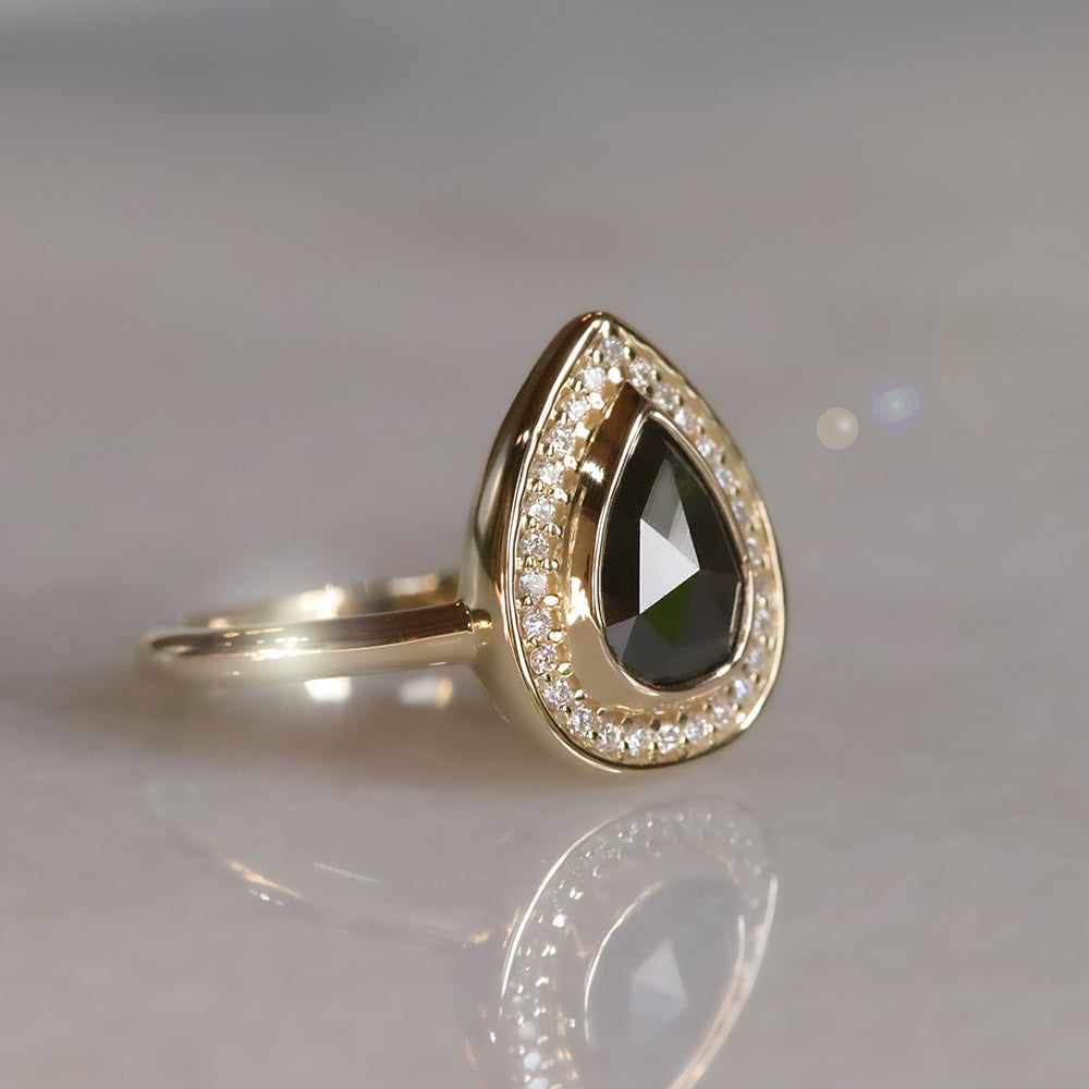 A side view of a pear-shaped black diamond ring is set in gold, surrounded by a halo of small, sparkling white diamonds. The background a light polished marble surface. 