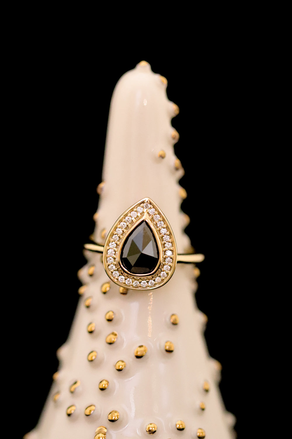 A pear-shaped black diamond ring is set in gold, surrounded by a halo of small, sparkling white diamonds. Displayed on a white and gold ceramic ring holder. 
