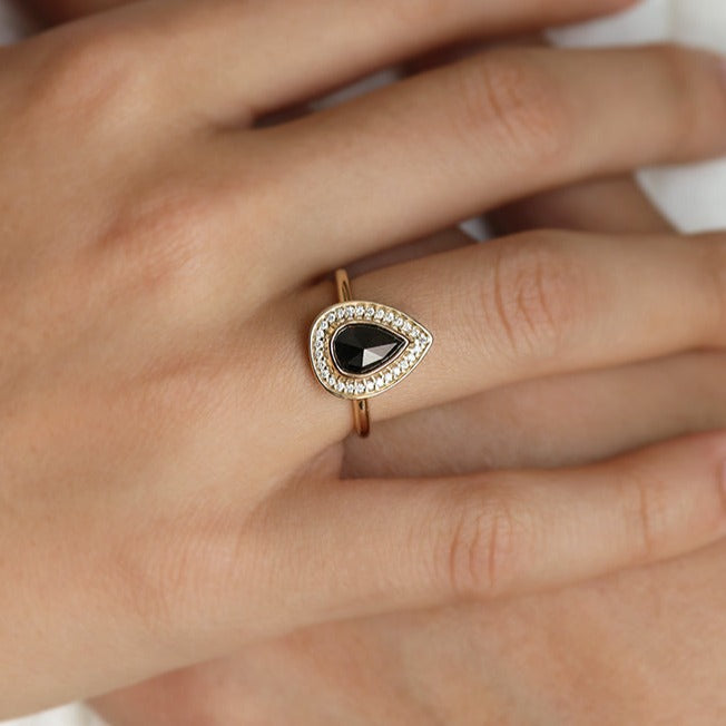 A pear-shaped black diamond ring is set in gold, surrounded by a halo of small, sparkling white diamonds. Worn on a hand.