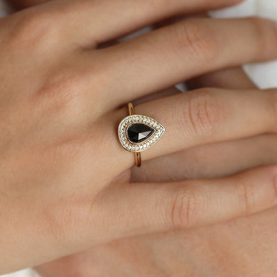 A pear-shaped black diamond ring is set in gold, surrounded by a halo of small, sparkling white diamonds. Worn on a hand.