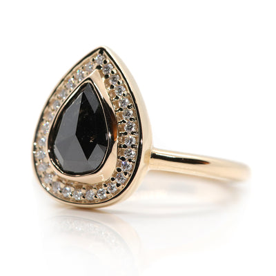 A pear-shaped black diamond ring is set in gold, surrounded by a halo of small, sparkling white diamonds. On a white background. 