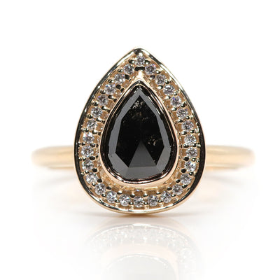 A pear-shaped black diamond ring is set in gold, surrounded by a halo of small, sparkling white diamonds. On a white background. 