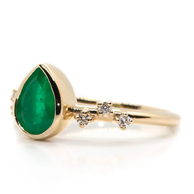 A pear-shaped emerald set in a gold bezel ring, adorned with offset small diamonds on the band. Displayed on a white background.