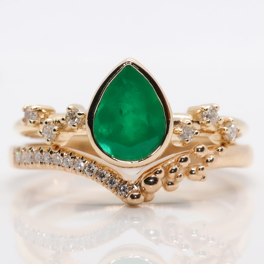 A pear-shaped emerald set in a gold bezel ring, adorned with offset small diamonds on the band. Stacked with an intricate crown-like diamond band. Displayed on a white background.